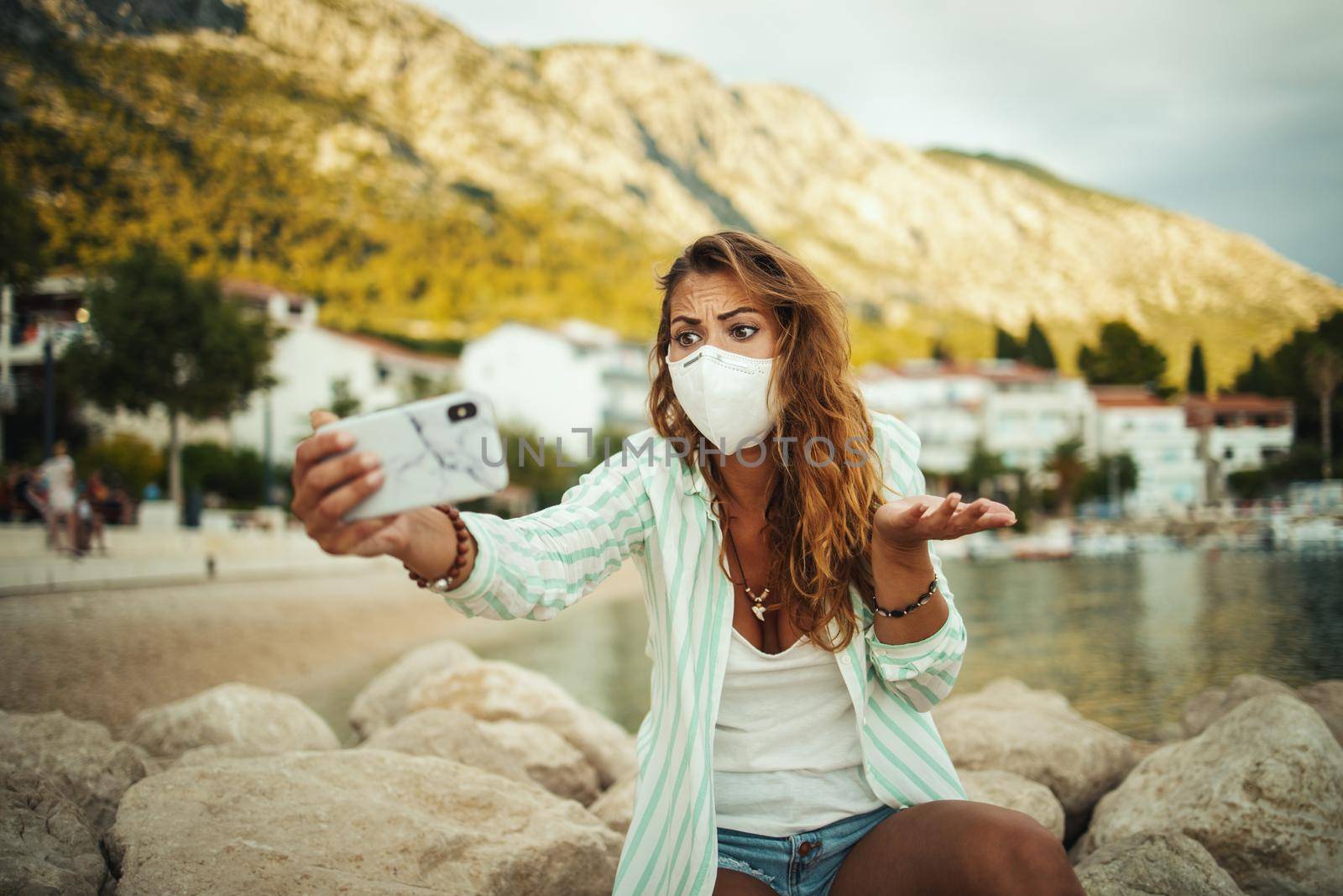 Shot of a concerned young woman wearing a protective N95 mask and making video call with her smartphone while spending time on the seaside during the COVID-19.