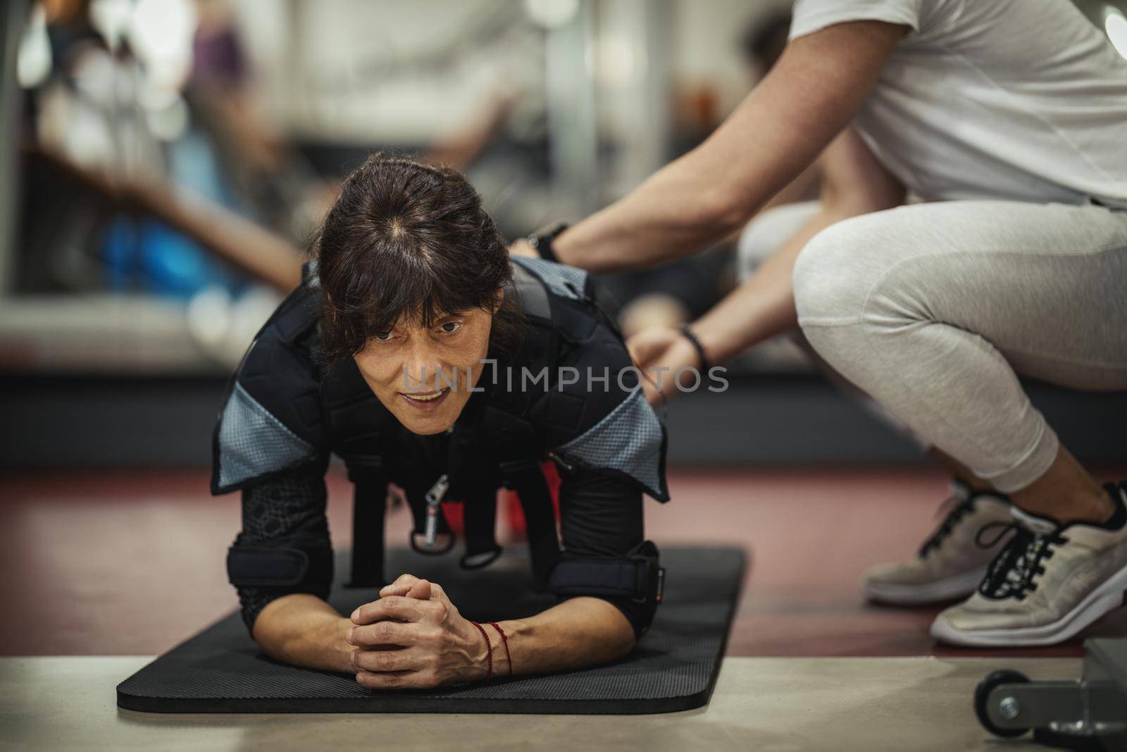 A smiling senior woman is doing plank exercises with personal trainer in the gym, dressed in a black suit with an EMS electronic simulator to stimulate their muscles.