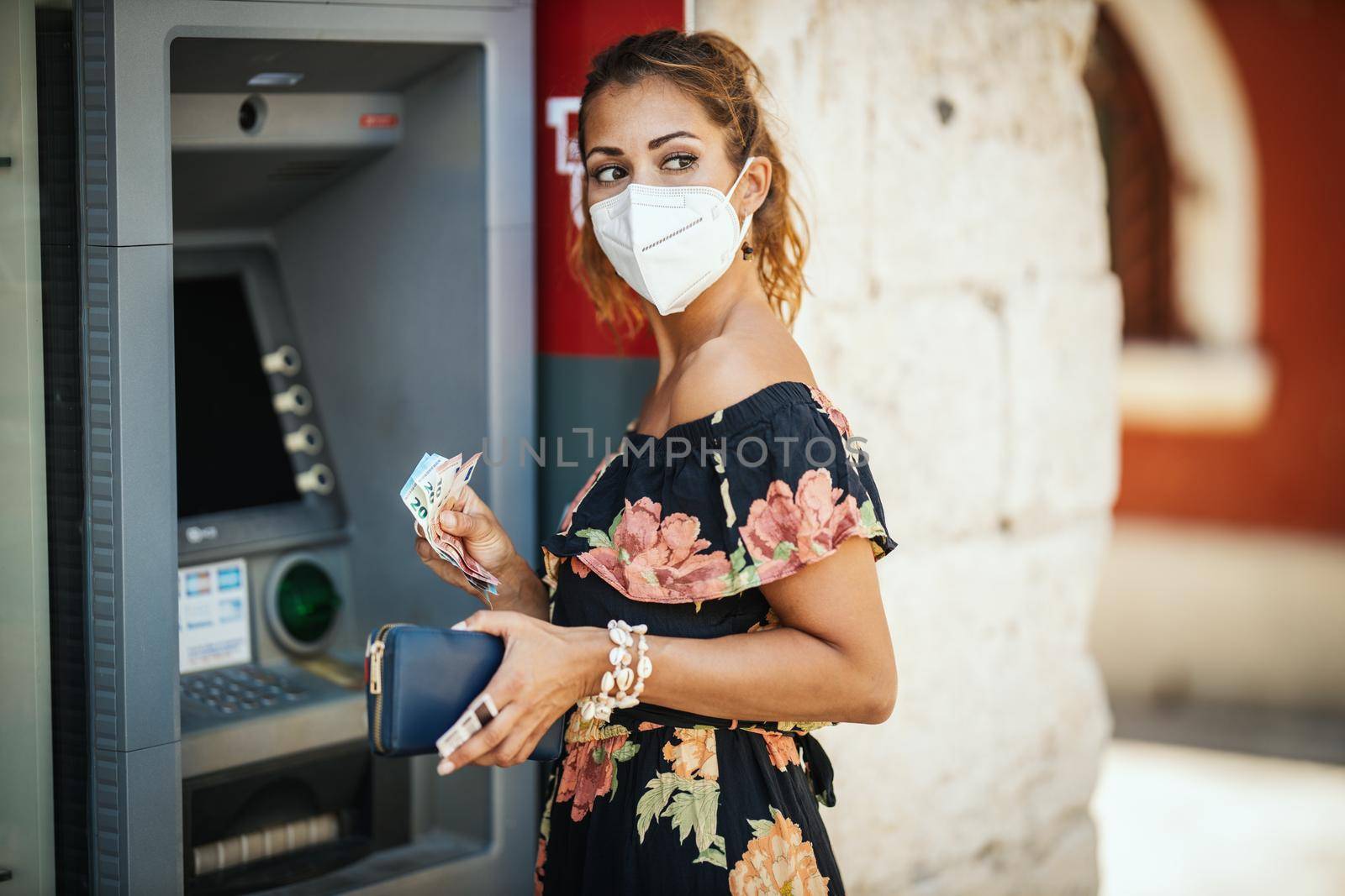 Side view of a young woman is wearing N95 protective mask while taking money from ATM machine during Covid-19 pandemic.