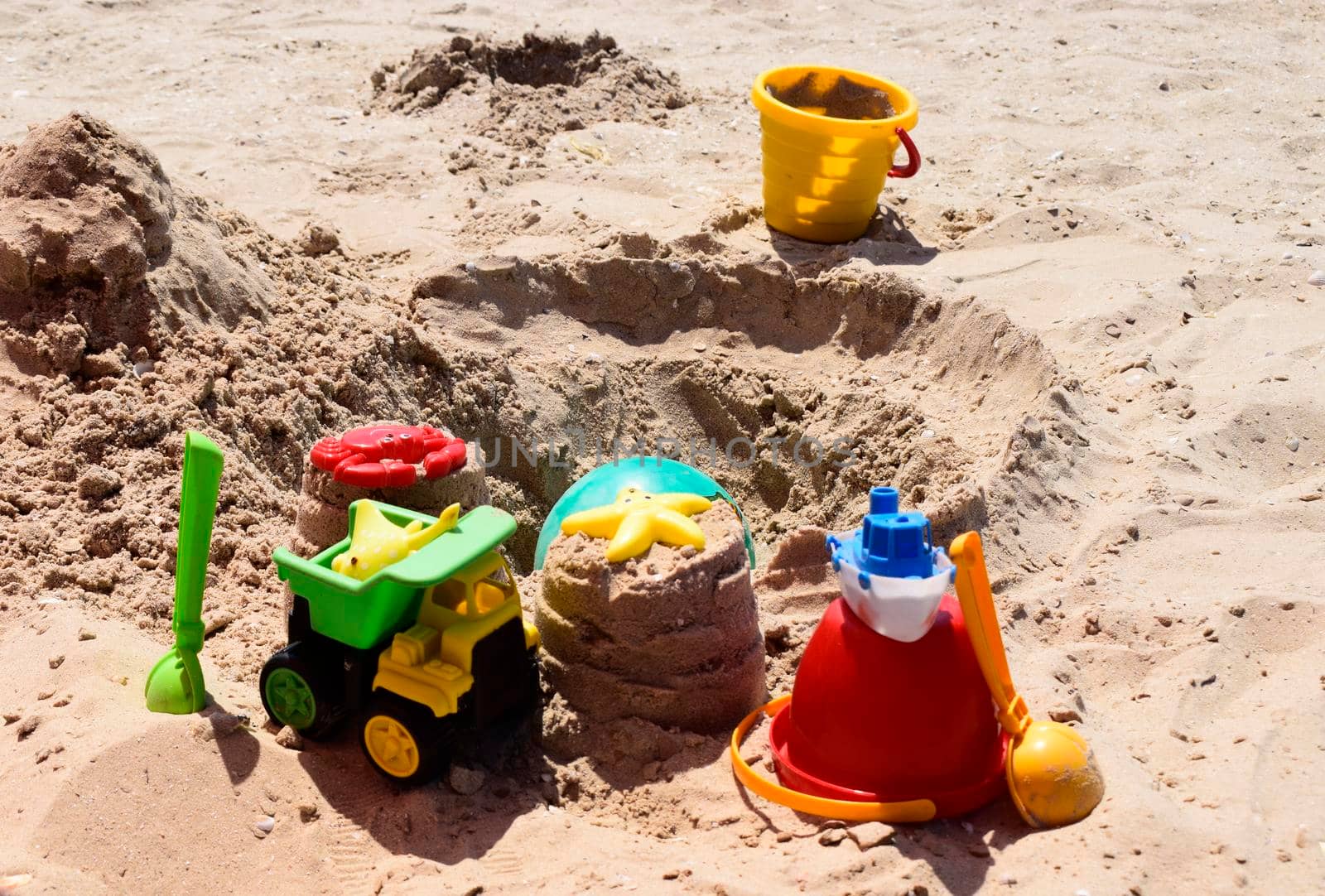 Children's plastic toys green end yellow car, shovel, yellow and red buckets, green ball with yellow sand on the beach by sea. Children's beach toys on sand on a sunny day. Sandbox on the playground