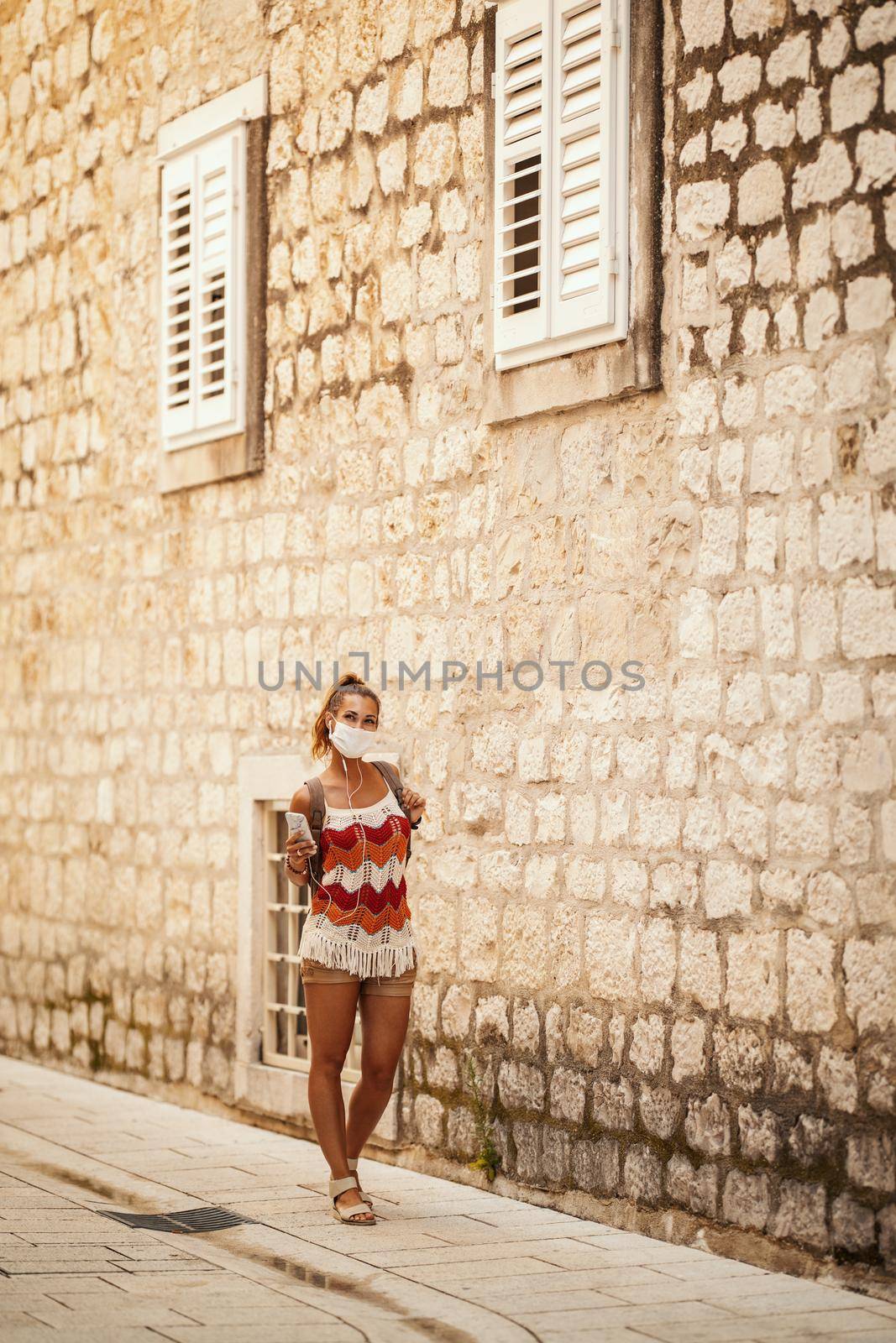 Shot of a happy young woman with protective mask spending time on vacation and exploring a Mediterranean city at corona pandemic. 