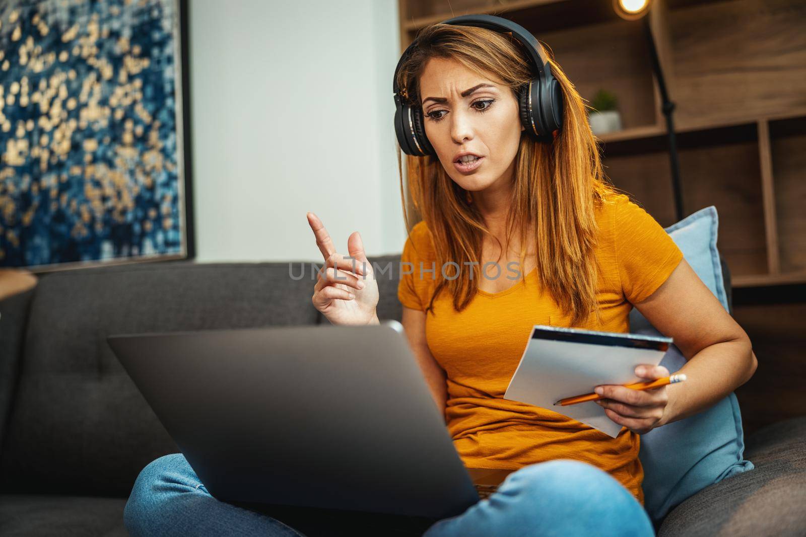 Cute young female student sitting on the sofa, using her laptop and headphones to watching lesson online and learning from home. Young woman taking notes while following teacher doing lesson on video call. 