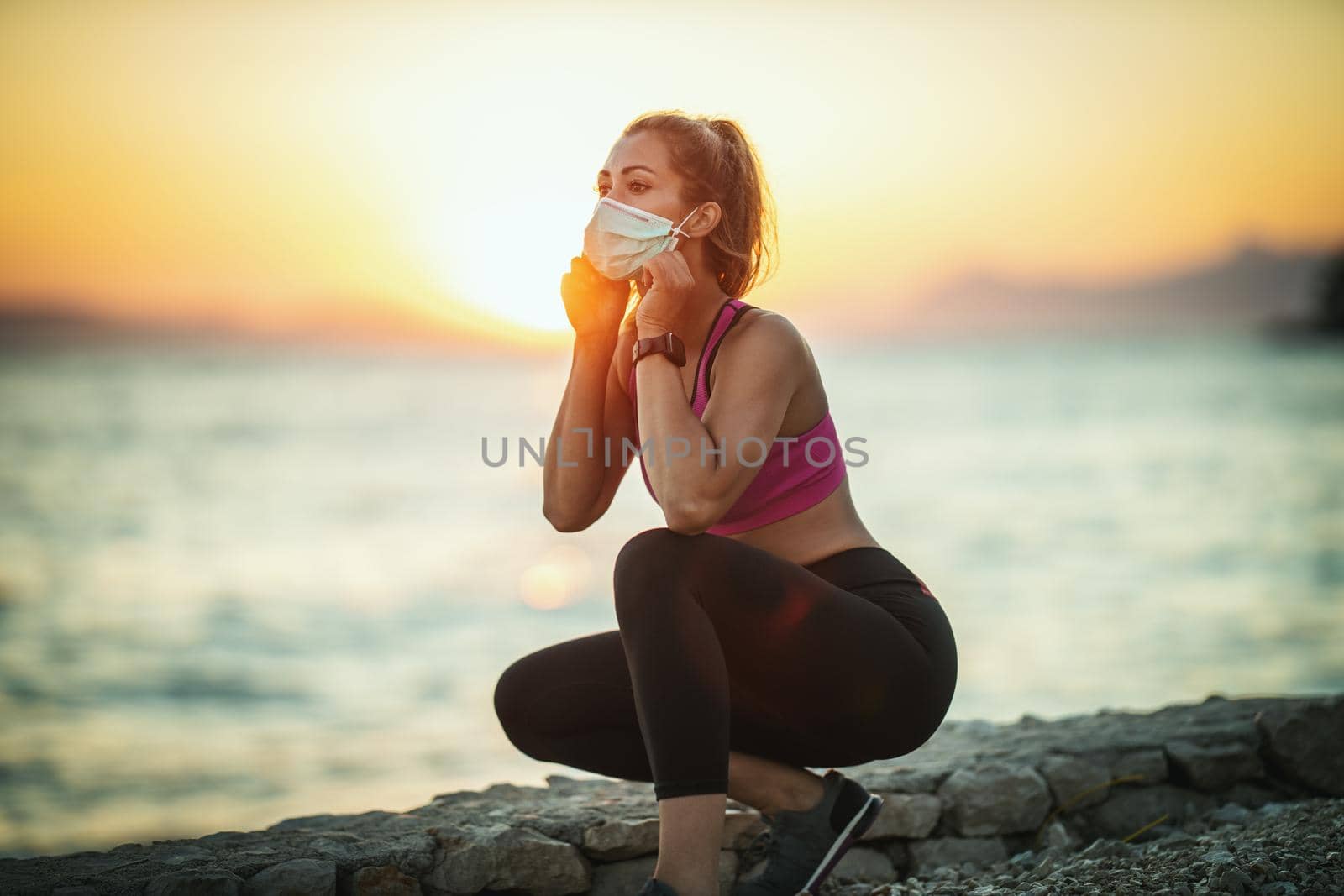 Shot of a female runner wearing protective face mask while doing training near the beach at sunset during the COVID-19 pandemic.