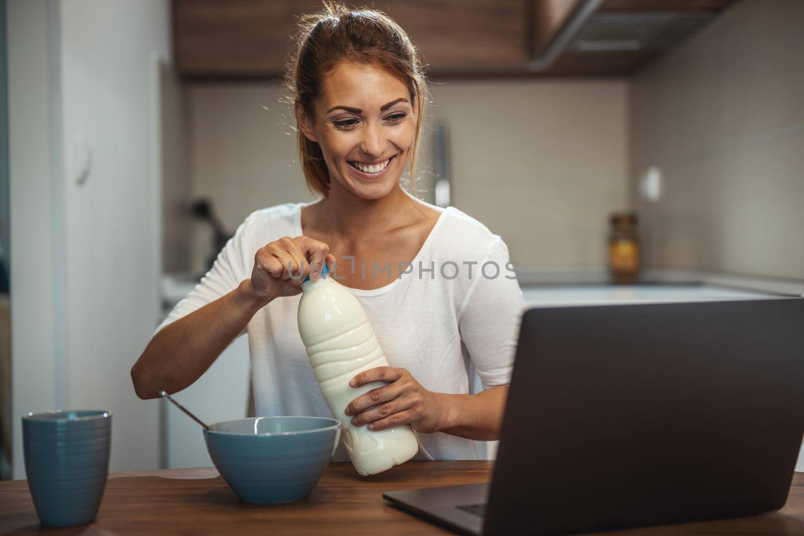 Beautiful young woman is preparing her healthy breakfast in her kitchen and using laptop to make a video chat with someone.