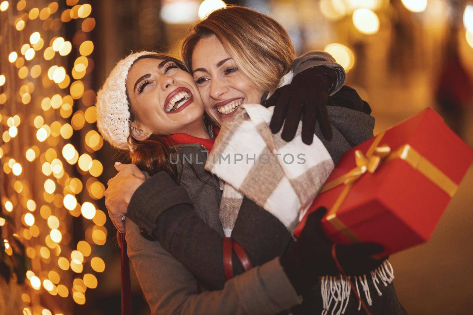 Cheerful two young women are having fun in the city street at the Christmas night. They are hugging and giving holidays presents to each other.