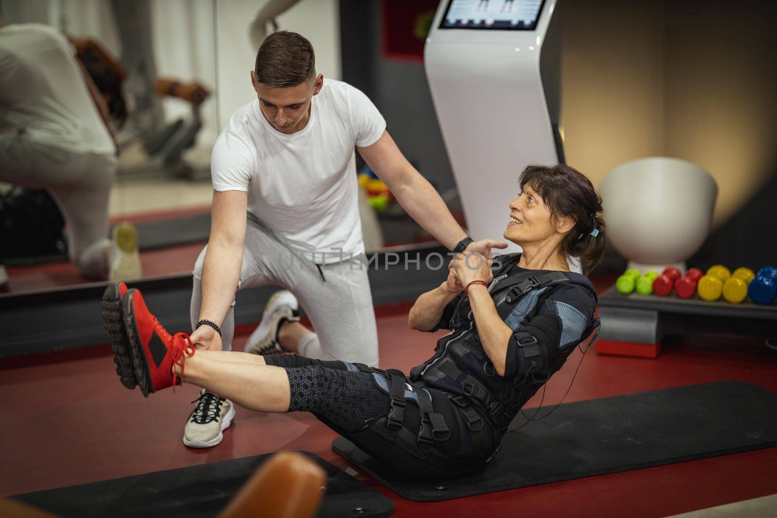 A smiling senior woman is doing sit-up exercises with personal trainer in the gym, dressed in a black suit with an EMS electronic simulator to stimulate their muscles.