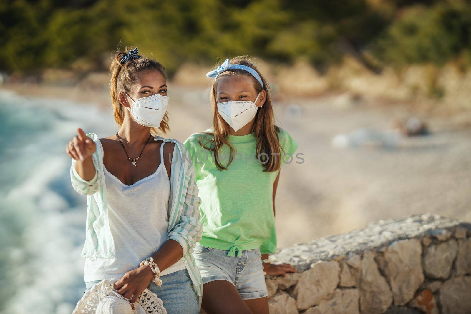 Shot of a smiling sisters with protective N95 mask spending time on the seashore at corona pandemic. They are having self-isolation in nature.
