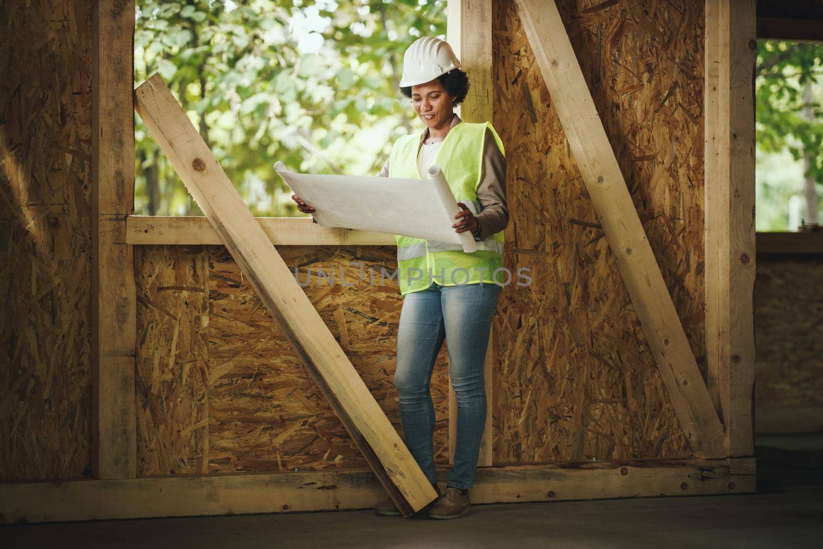 Shot of an African female architect checking plans at the construction site of a new wooden house. She is wearing protective workwear and white helmet.