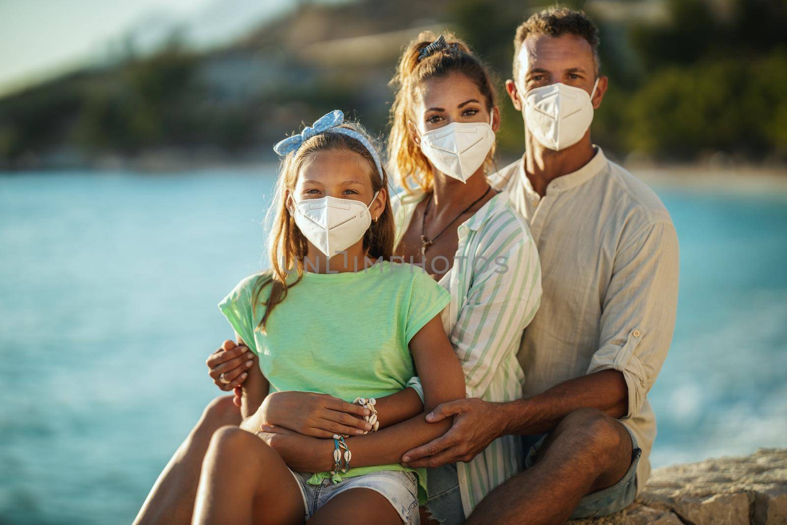 Portrait of a happy embraced family with protective N95 mask spending time on the seaside at corona pandemic. 