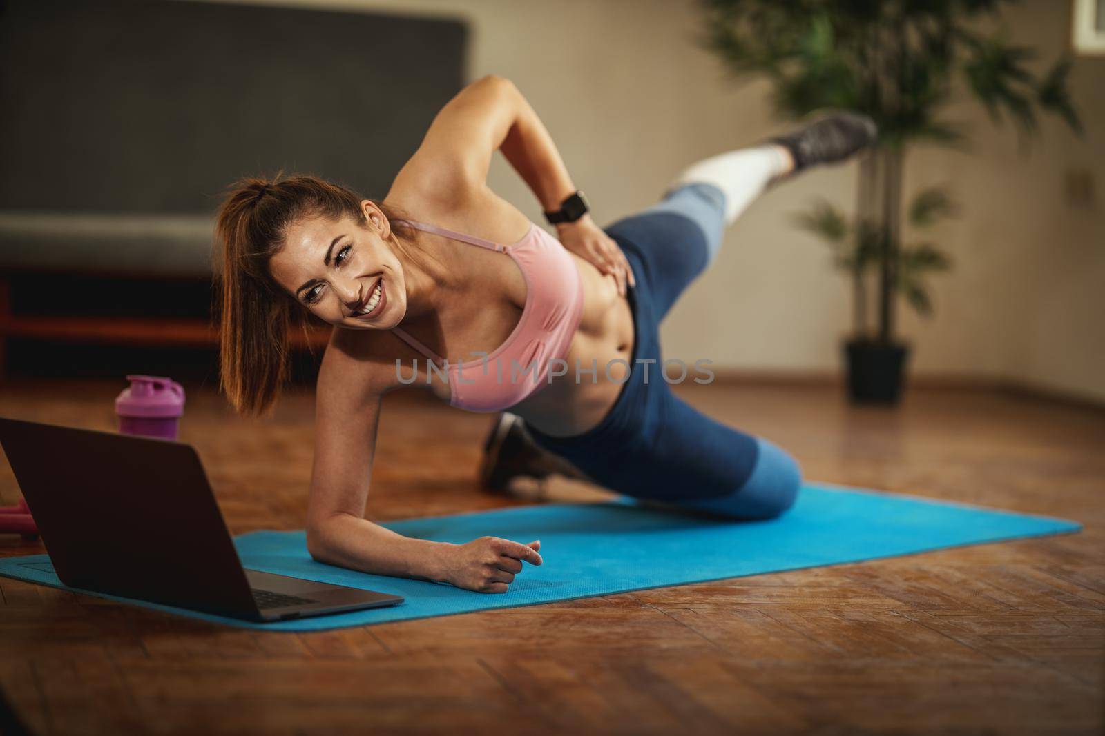 Young smiling woman is doing fitness exercises in the living room on floor mat and looking at the laptop, in morning sunshine.