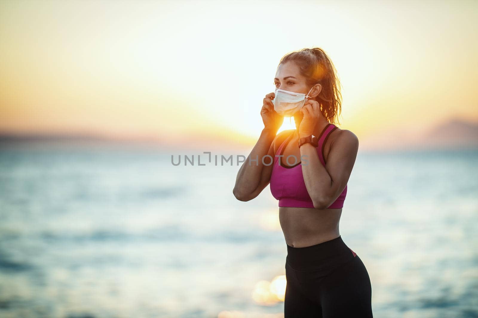 Shot of a female runner wearing protective face mask while doing training near the beach at sunset during the COVID-19 pandemic.