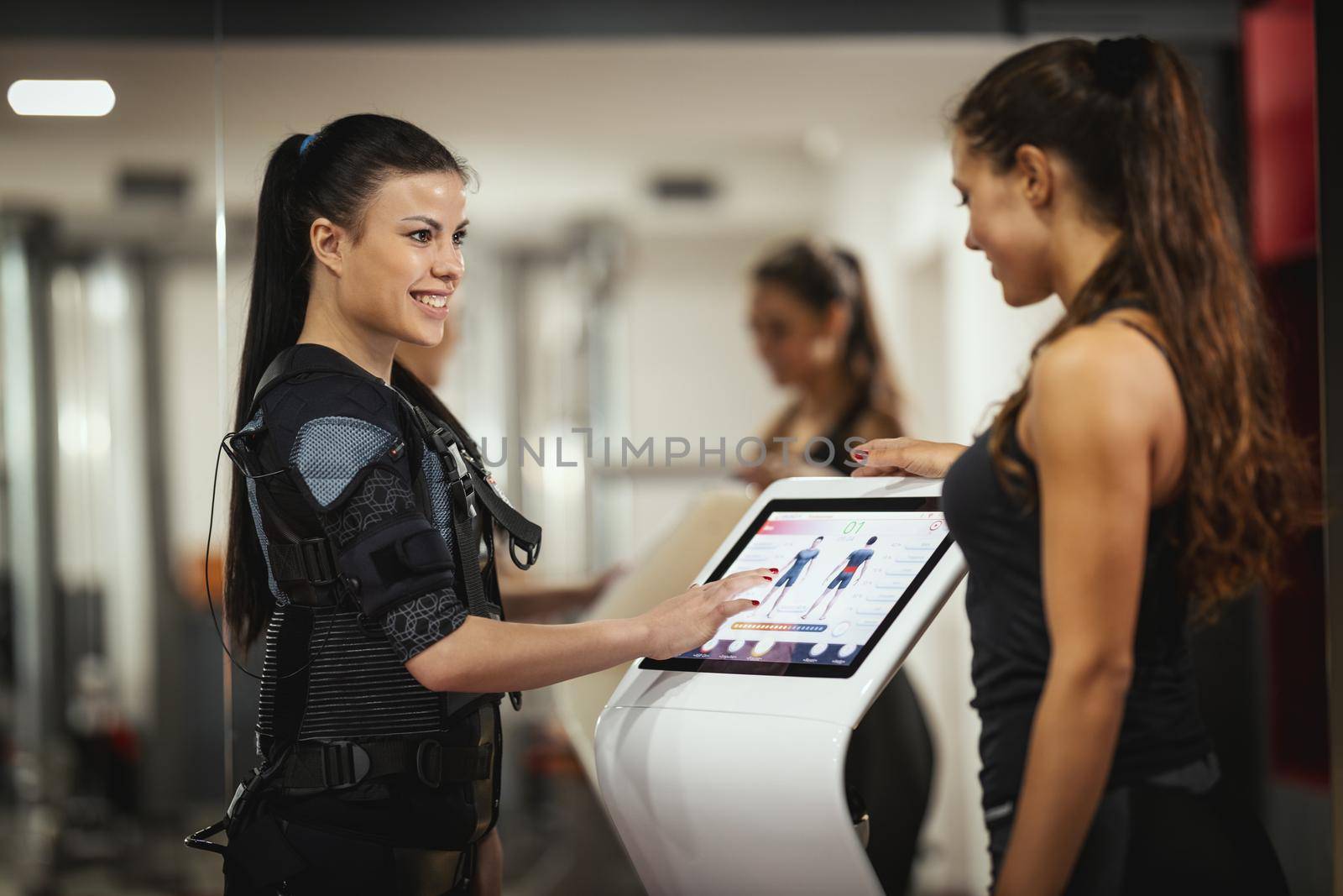 Female personal trainer is teaching a smiling young woman to workout with EMS device in the gym.