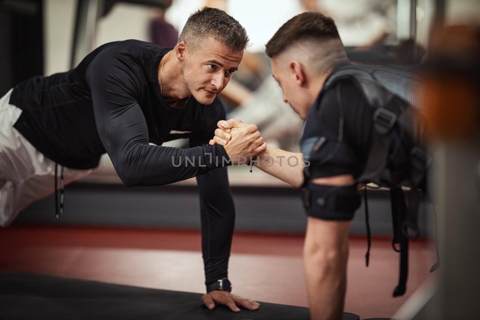 A young man is doing plank exercises with personal trainer in the gym, dressed in a black suit with an EMS electronic simulator to stimulate his muscles.