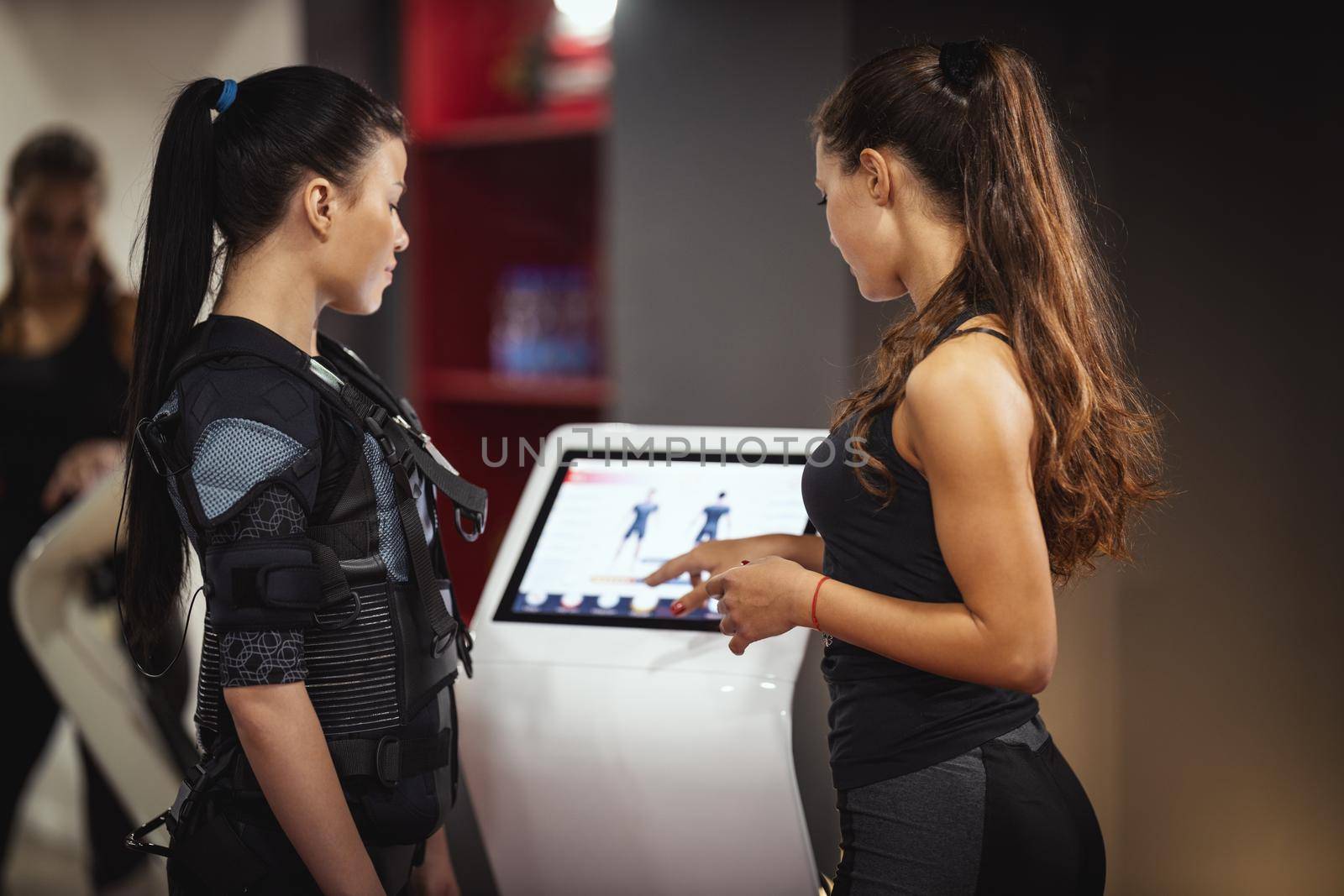Female personal trainer is teaching a smiling young woman to workout with EMS device in the gym.