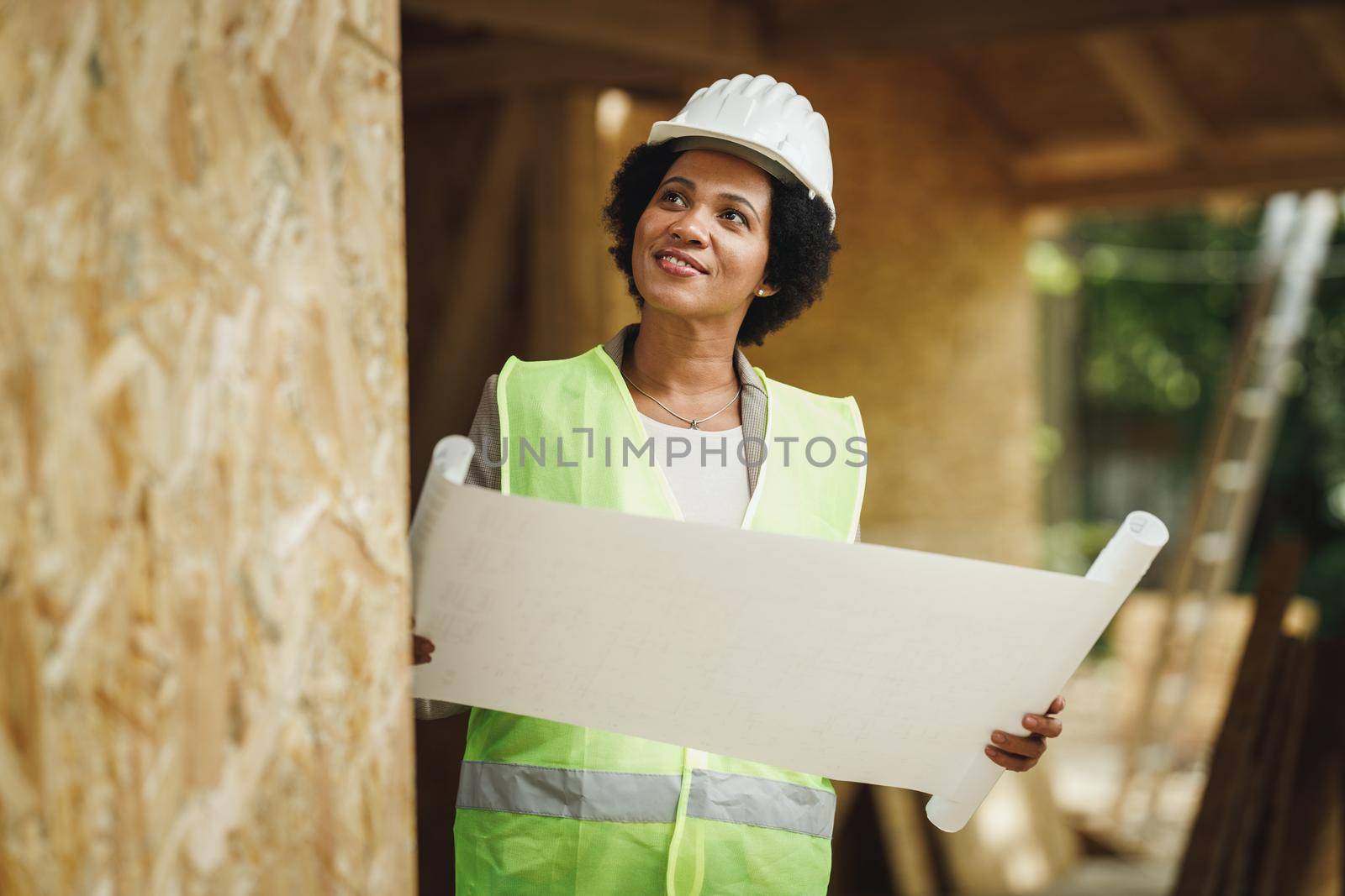 Shot of an African female architect checking blueprints at the construction site of a new wooden house. She is wearing protective workwear and white helmet.