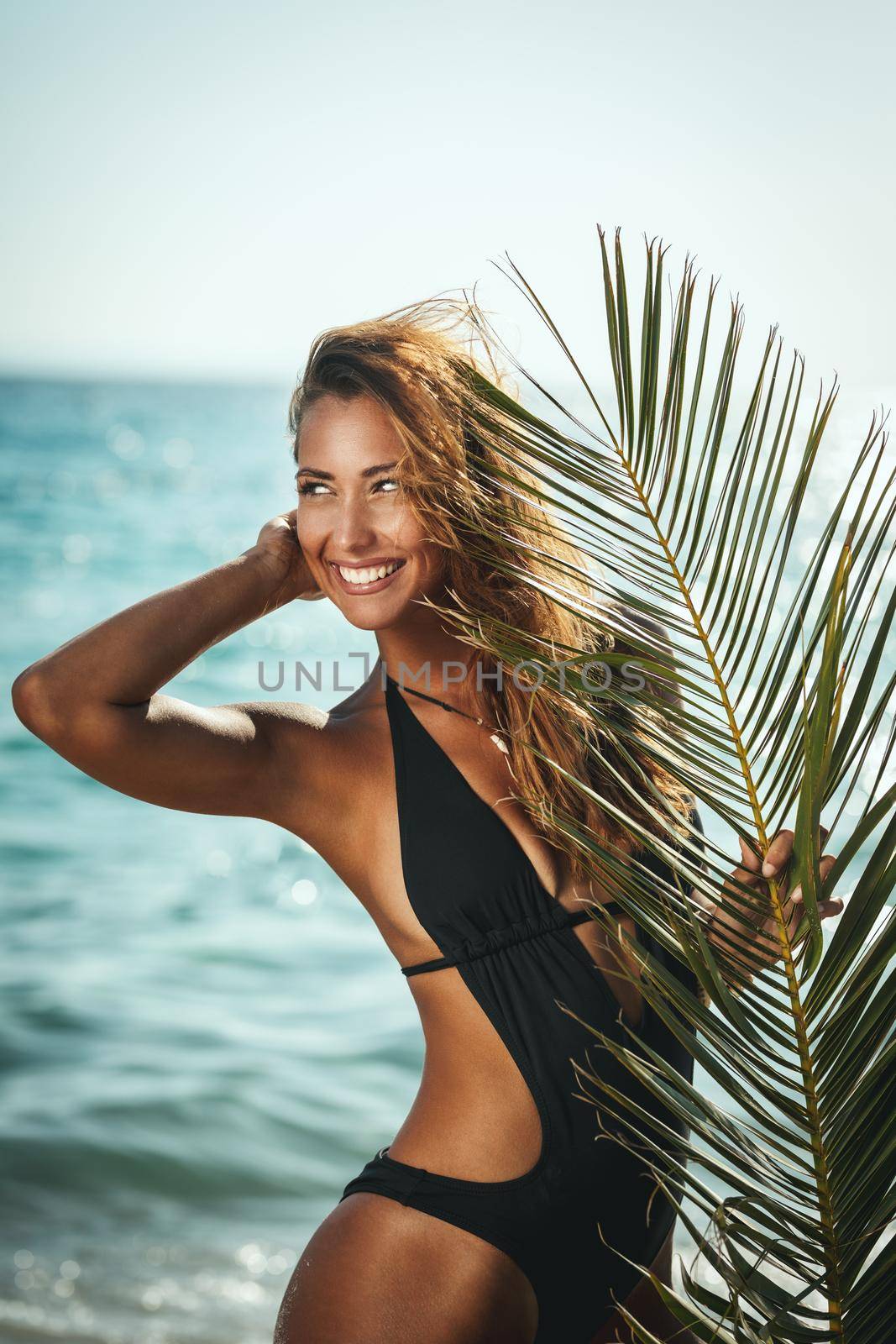 Attractive young woman is posing and enjoying covered with palm tree leaf at the tropical beach.