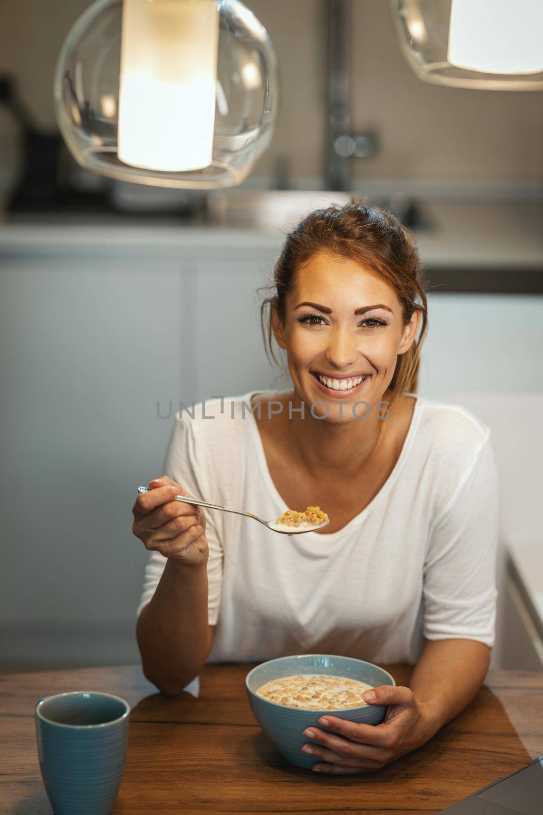 Beautiful young smiling woman is eating healthy breakfast in her kitchen. Looking at camera.