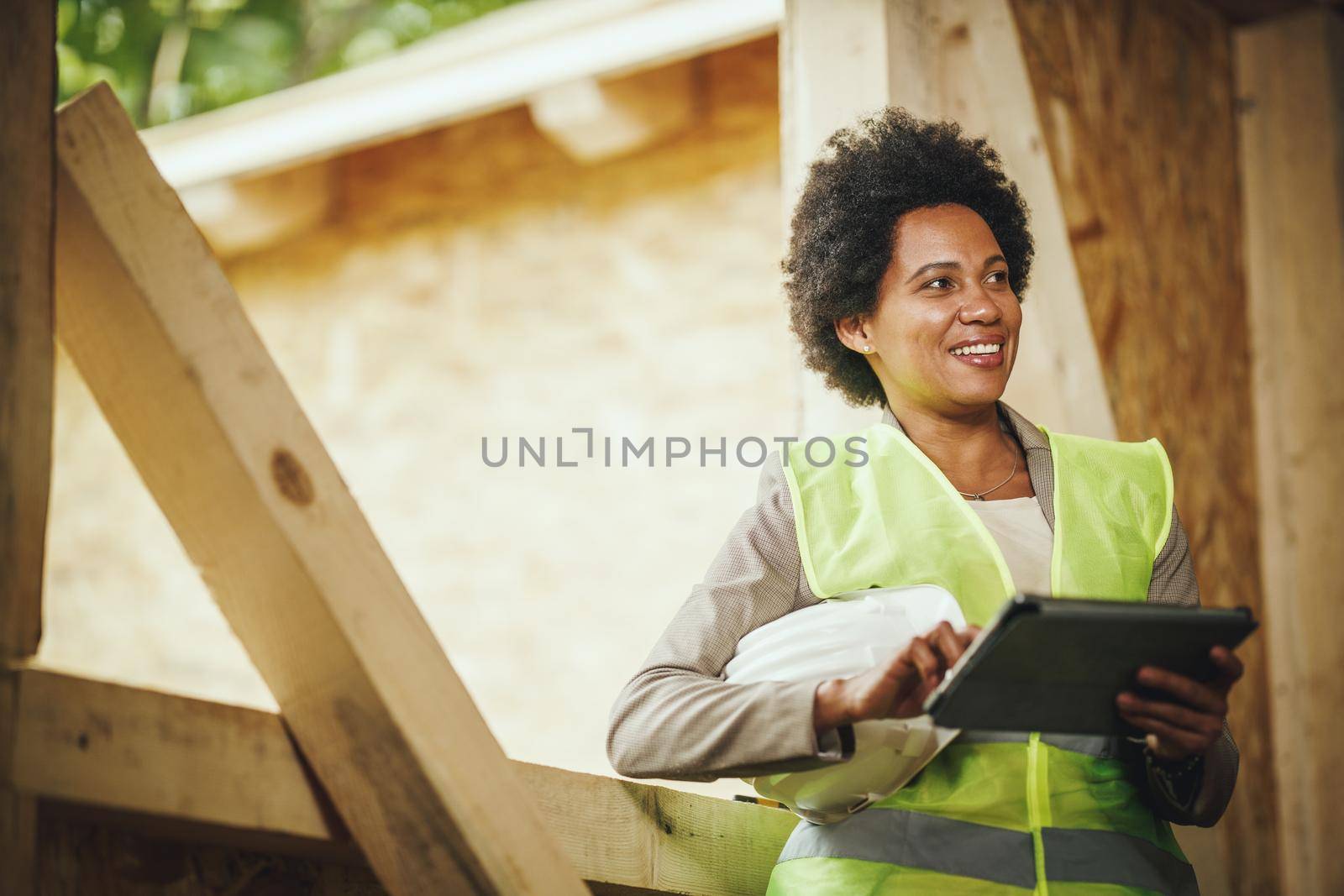 Shot of an African female architect using a digital tablet at the construction site of a new wooden house. She is wearing protective workwear and white helmet.
