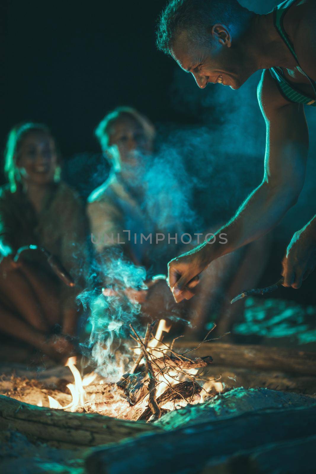 The happy young smiling friends are sitting on the sandy seashore by the fire in the evening. They are frying sausages on stick.