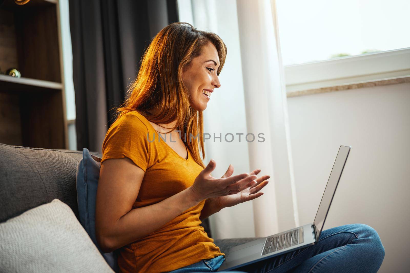Attractive young woman sitting cross legged on the sofa and using her laptop to make a video chat with someone at home.