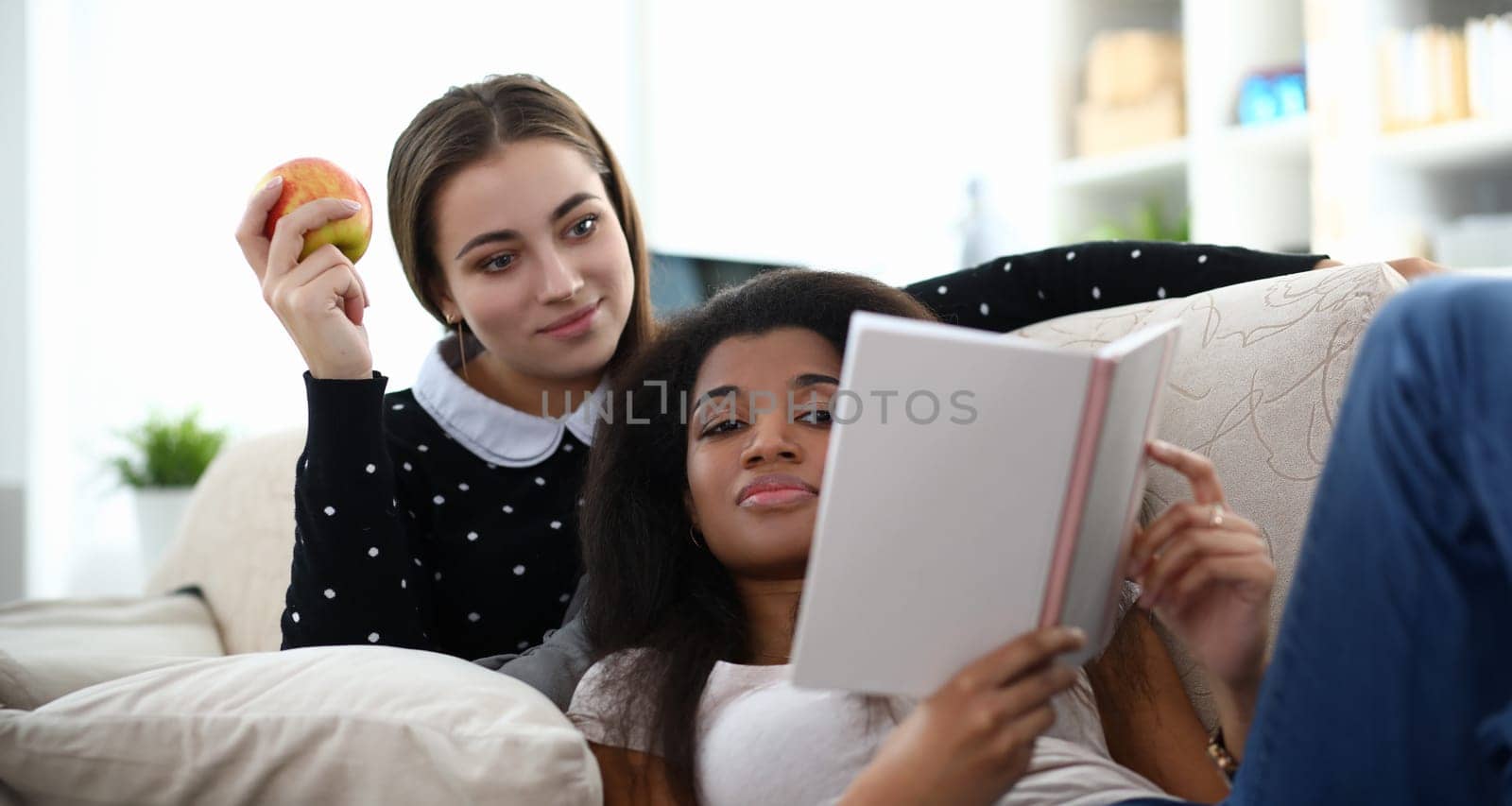 Portrait of best friends reading diary together on couch. Young beautiful woman with apple. Smiling female spending leisure time. Cozy home interior and relaxation concept