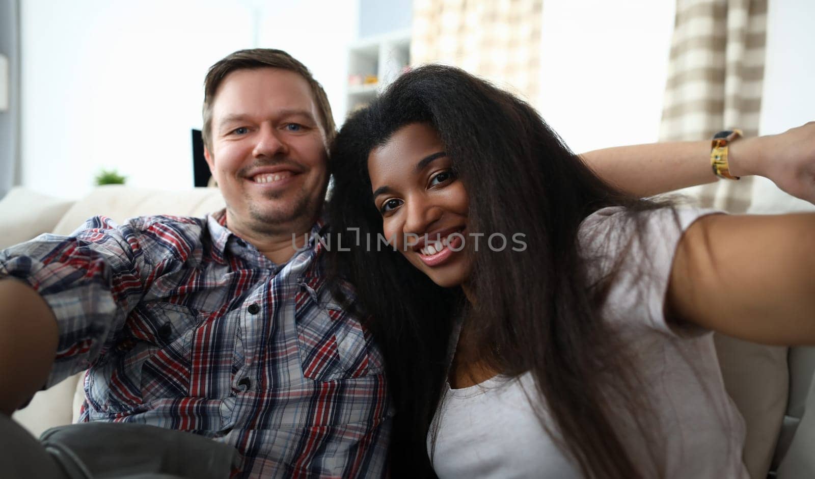 Portrait of cheerful mixed race couple taking happy selfie on sofa indoors. by kuprevich