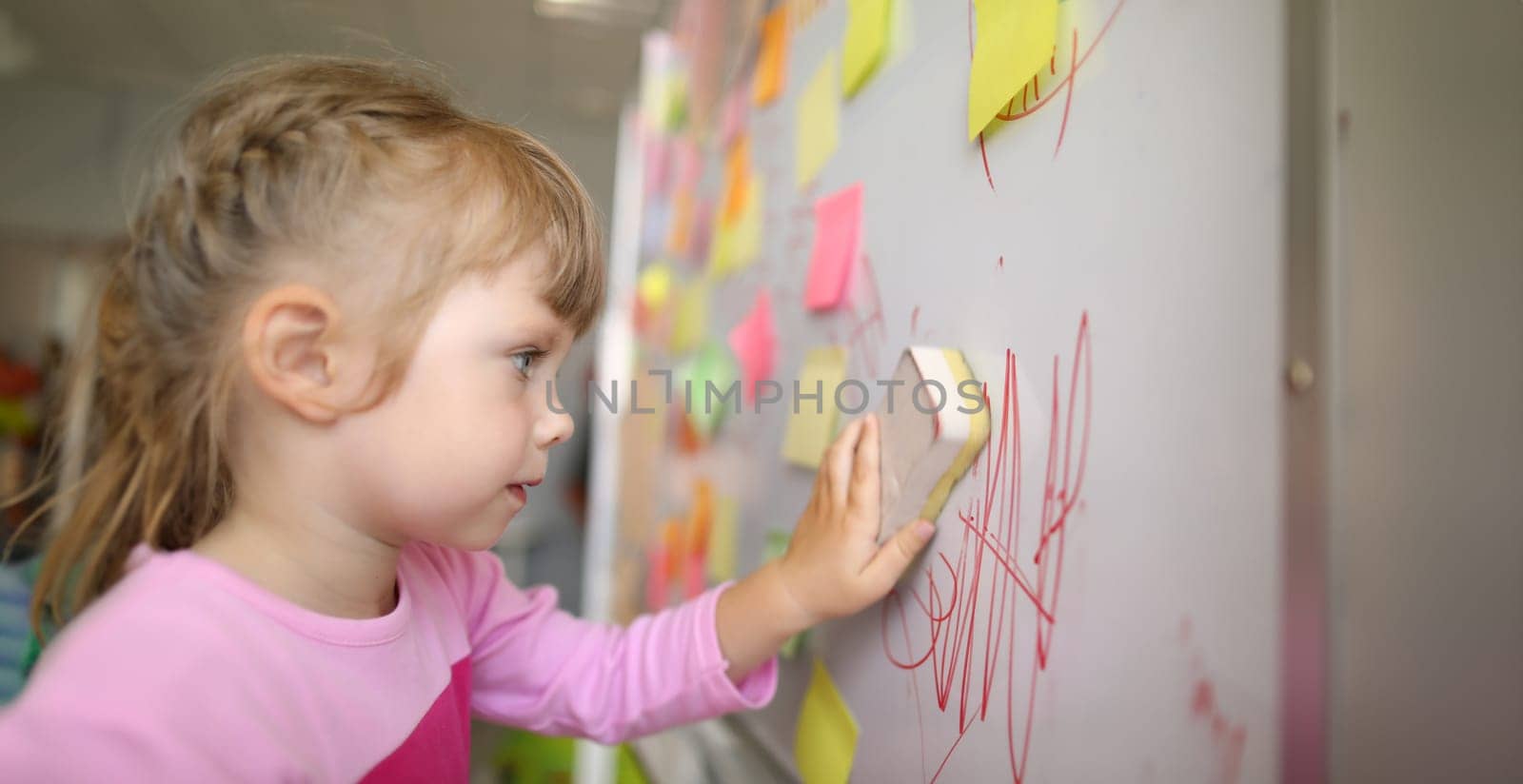 Close-up view of kid washes off the board in class with sponge. Child wiping doodle written on chalkboard. Many sticky notes on whiteboard. Childhood concept