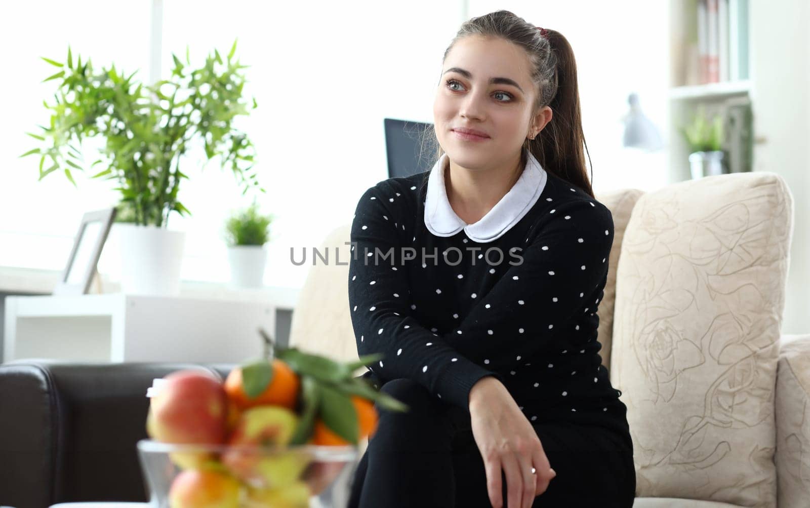 Portrait of smiling attractive young woman sitting on sofa in cozy interior. Lovely brunette smiling and looking away with gladness. Bowl with fruits on table