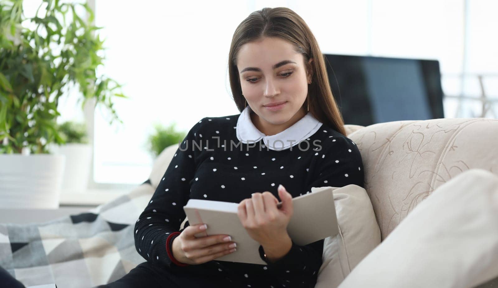 Portrait of young charming lady reading interesting book in living room. Smiling woman looking at notebook with gladness. Relaxation and spare time concept