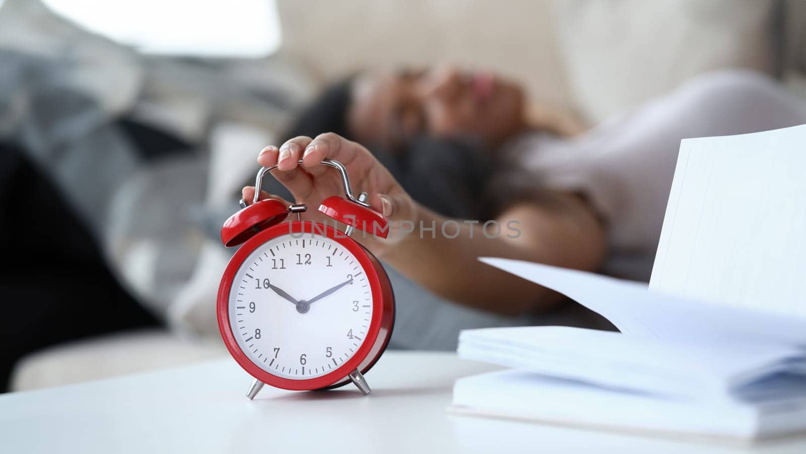 Close-up of bright red alarm clock and young latino woman turning it off while laying in bed. Ten minutes to eleven. Book pages on table near wake-up call