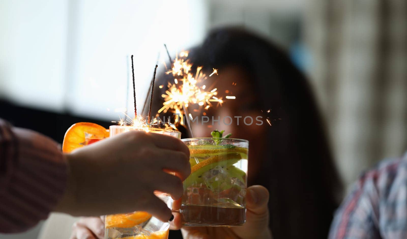 Close-up of friends hands holding fruit cocktails with sparklers decoration. Delicious refreshing beverage. Christmas, new year celebration party and holiday concept