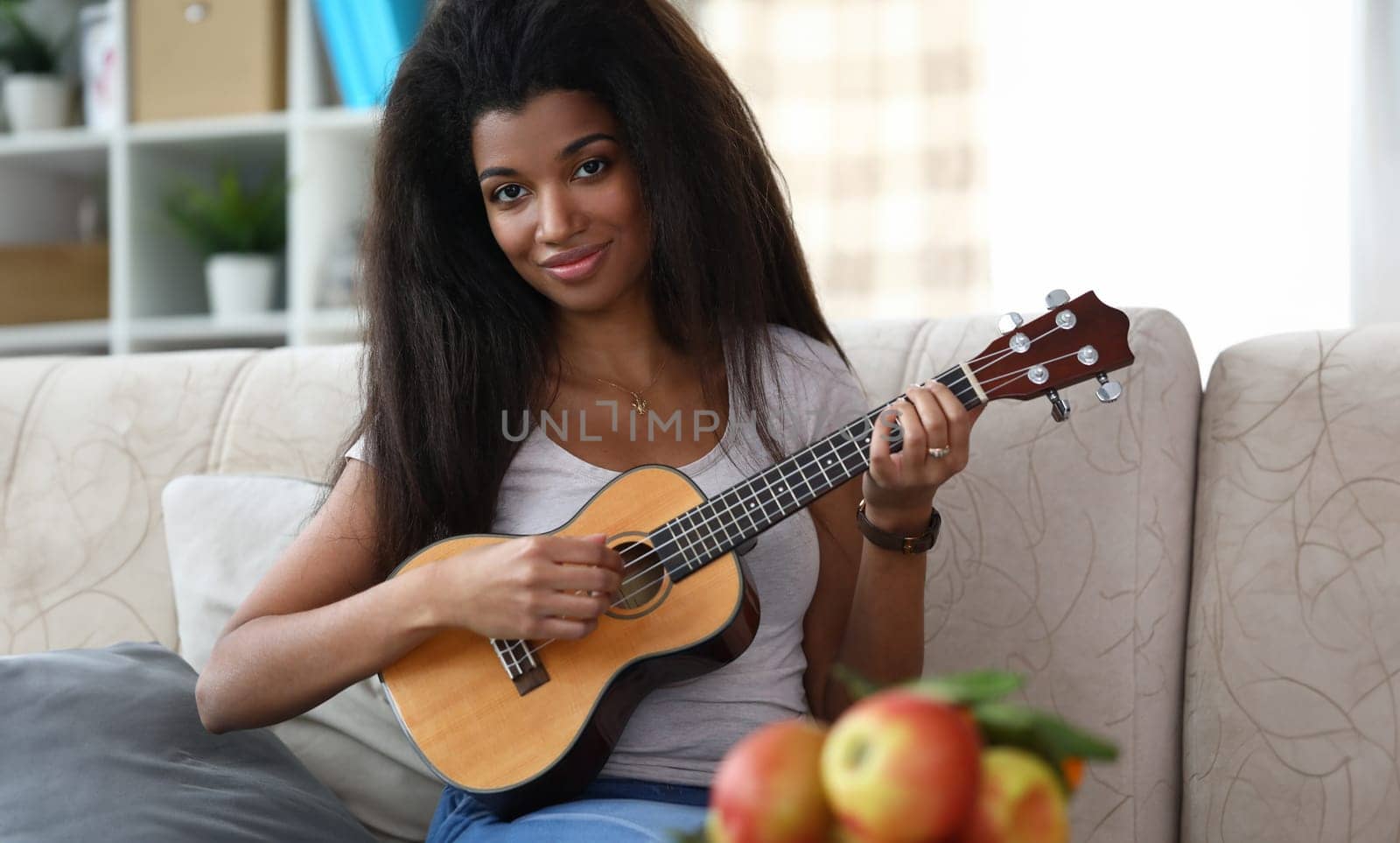 Portrait of smiling african-american woman playing on brown ukulele guitar. Latino musician sitting on comfy sofa. Cheerful lady looking at camera with happiness. Music and entertainment concept