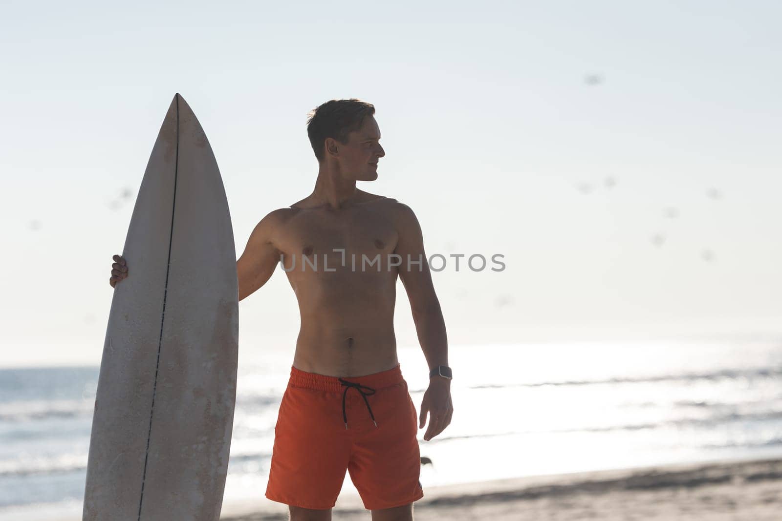 A shirtless man surfer standing on the beach holding a surfboard - looking to the side. Mid shot