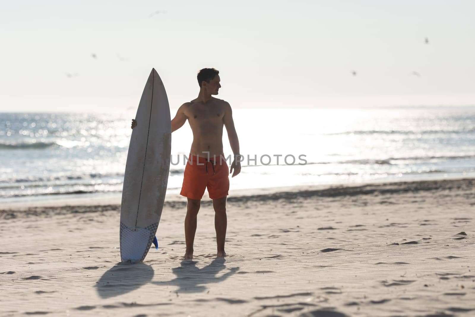 A shirtless man surfer standing on the beach holding a surfboard. Mid shot