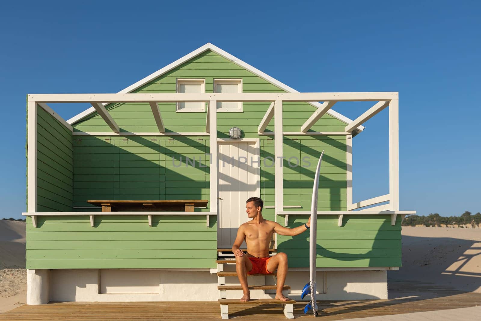 A man surfer sits on the stairs at a shore house on the beach. Mid shot