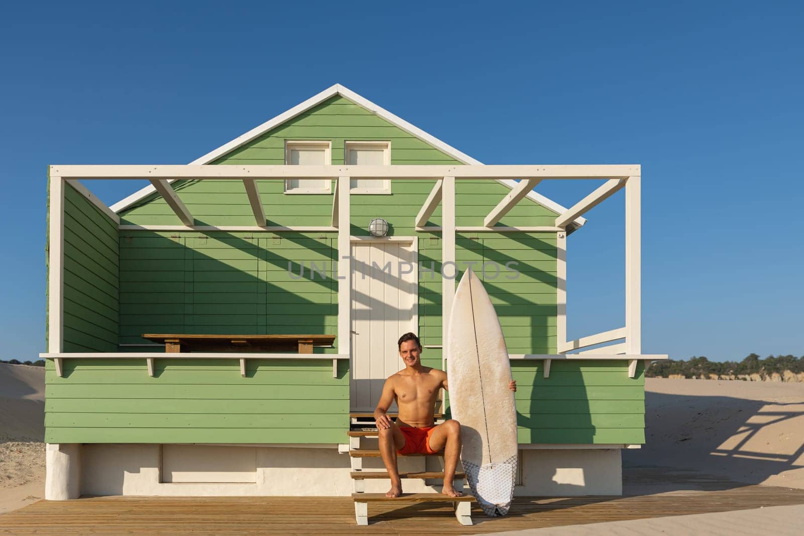 A smiling man surfer sits on the stairs at a shore house on the beach. Mid shot