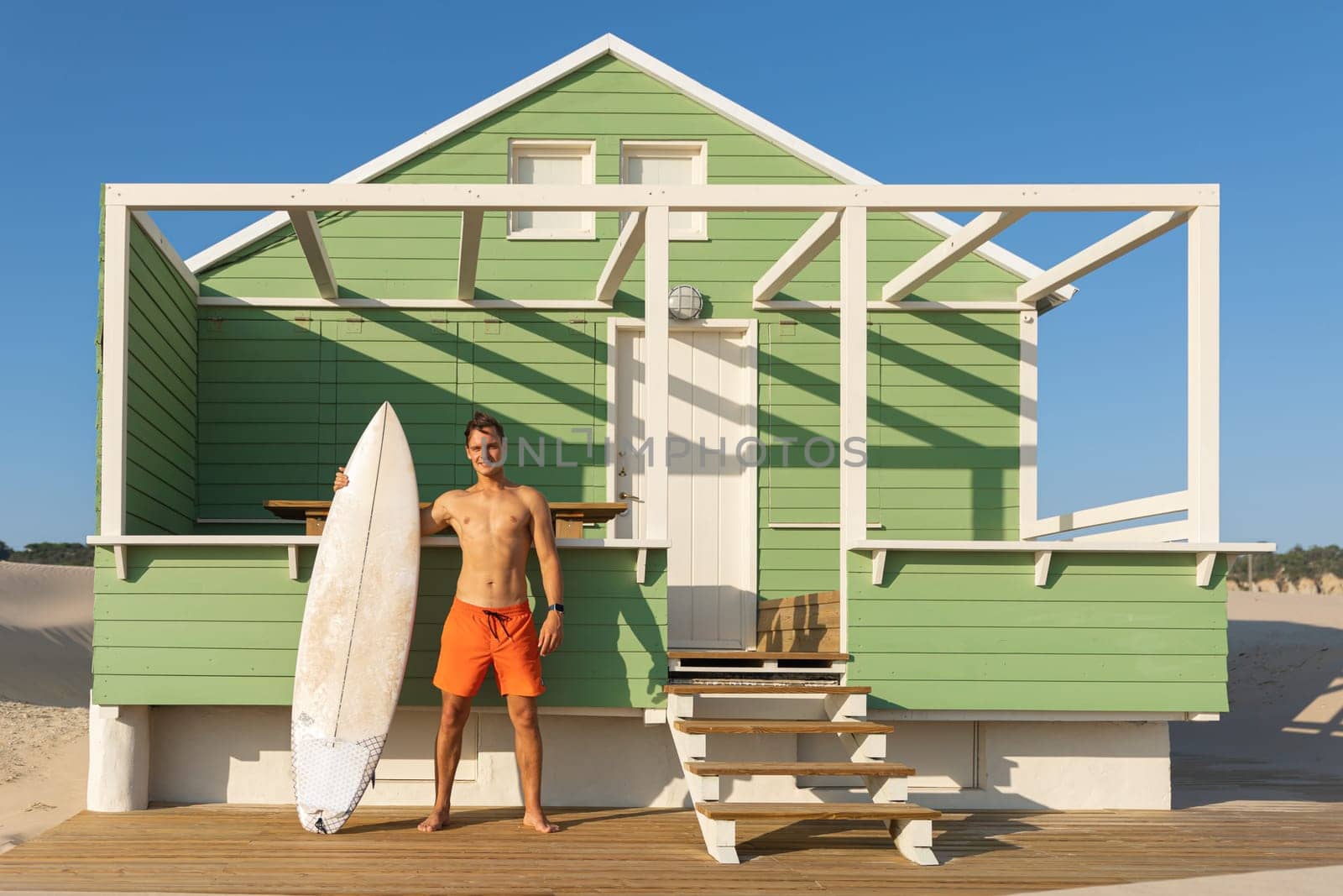 A smiling man surfer standing at a shore house on the beach holding his surfing board. Mid shot