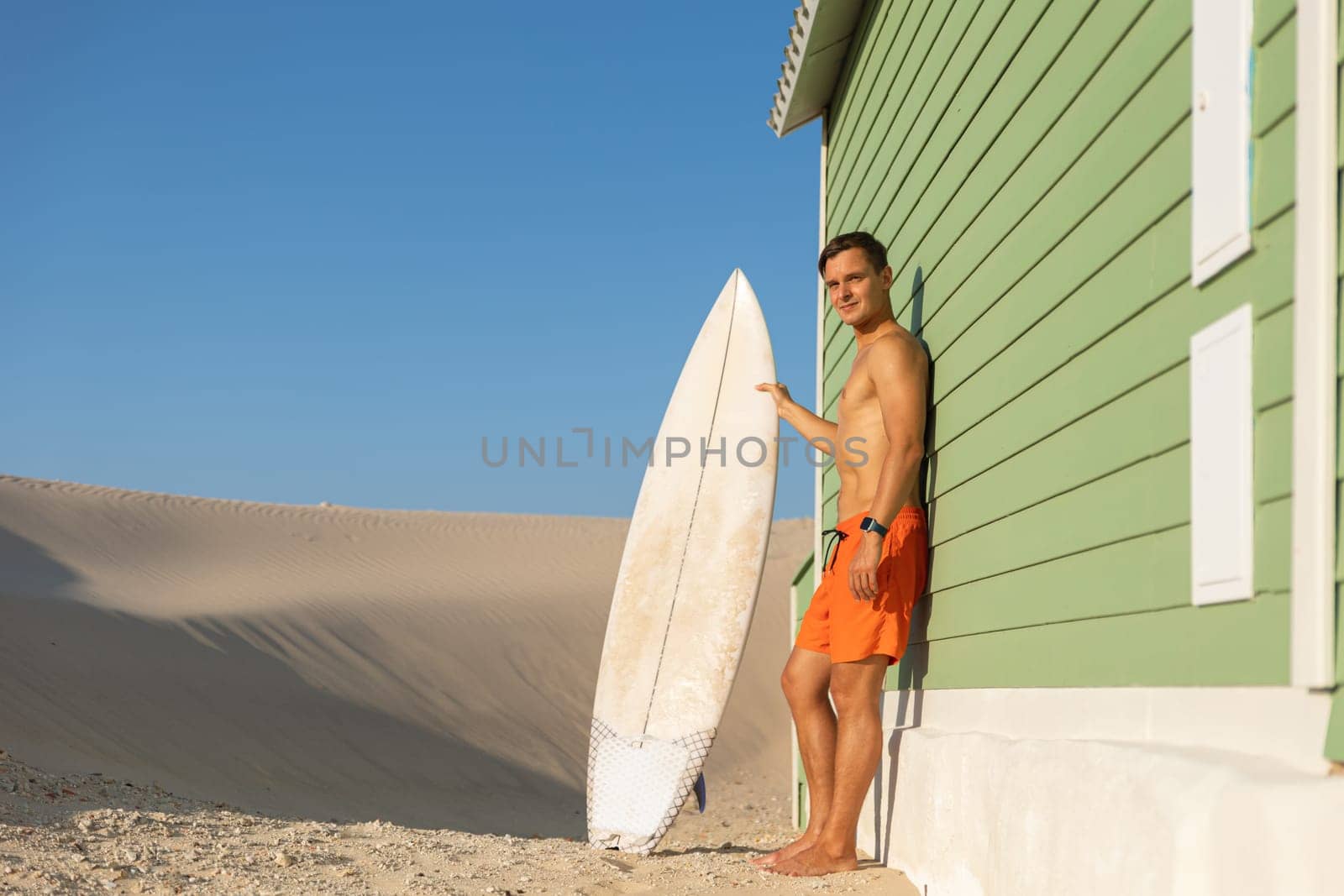 A man surfer standing at the wall of a shore house holding his surfing board. Mid shot