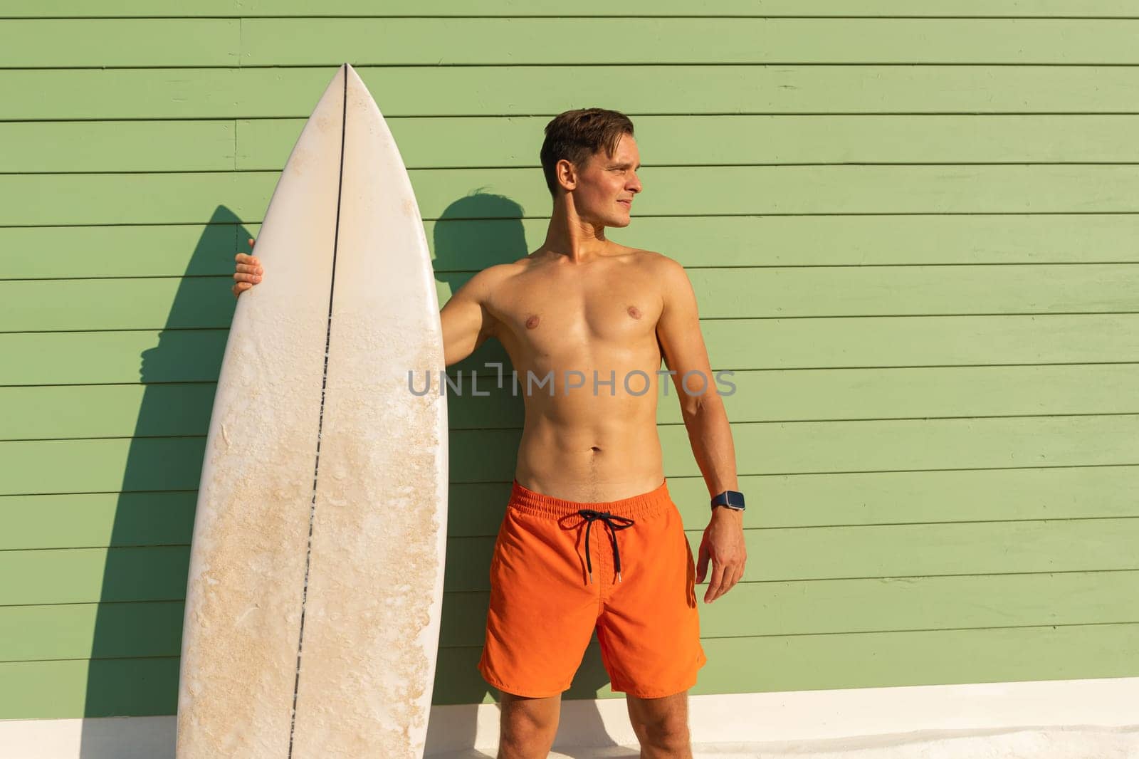 A man standing at the light green wall holding a surfboard. Mid shot