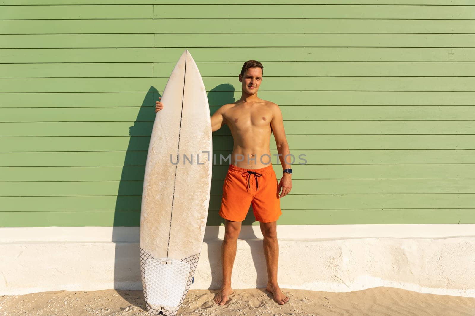 A man with nice body standing at the light green wall holding a surfboard. Mid shot