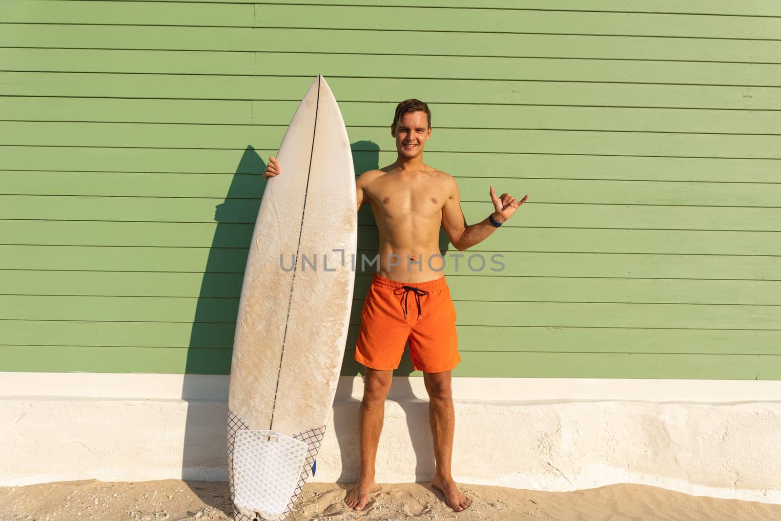 A smiling man with nice body standing at the light green wall holding a surfboard and showing shaka. Mid shot