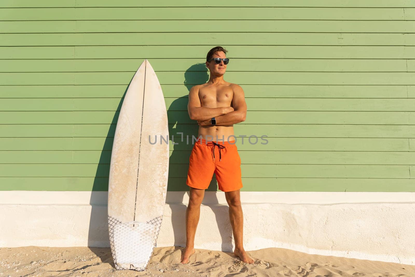 A smiling man with nice body wearing sunglasses standing at the light green with a surfboard. Mid shot