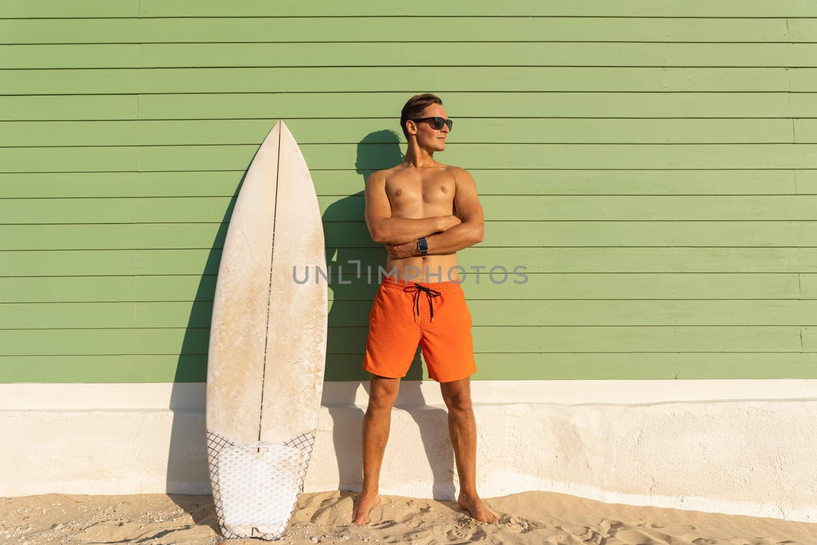 A smiling man with nice body wearing sunglasses standing at the light green with a surfboard - looking to the side. Mid shot