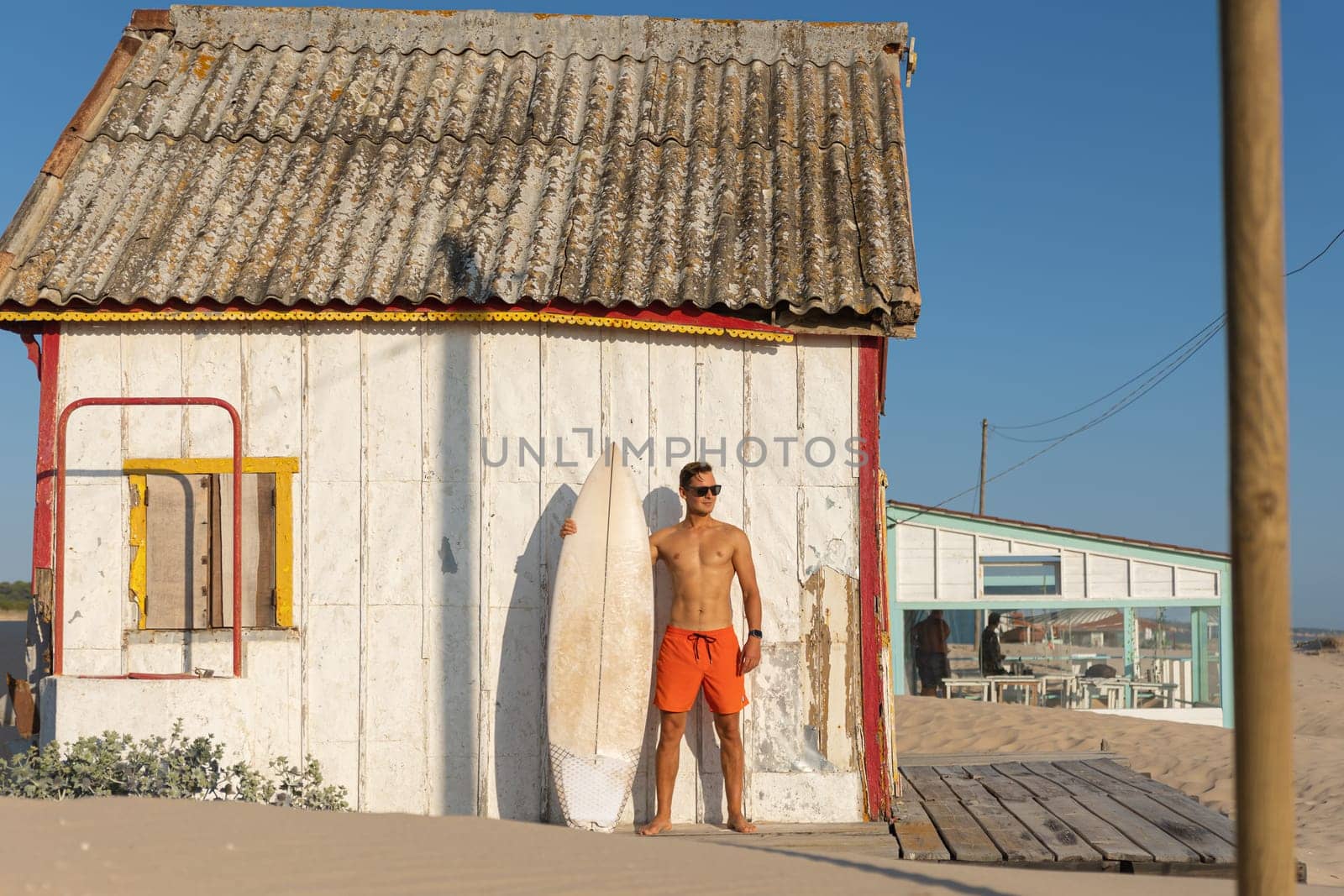 A man with nice athletic body standing at the shore house on the beach with his surfboard. Mid shot