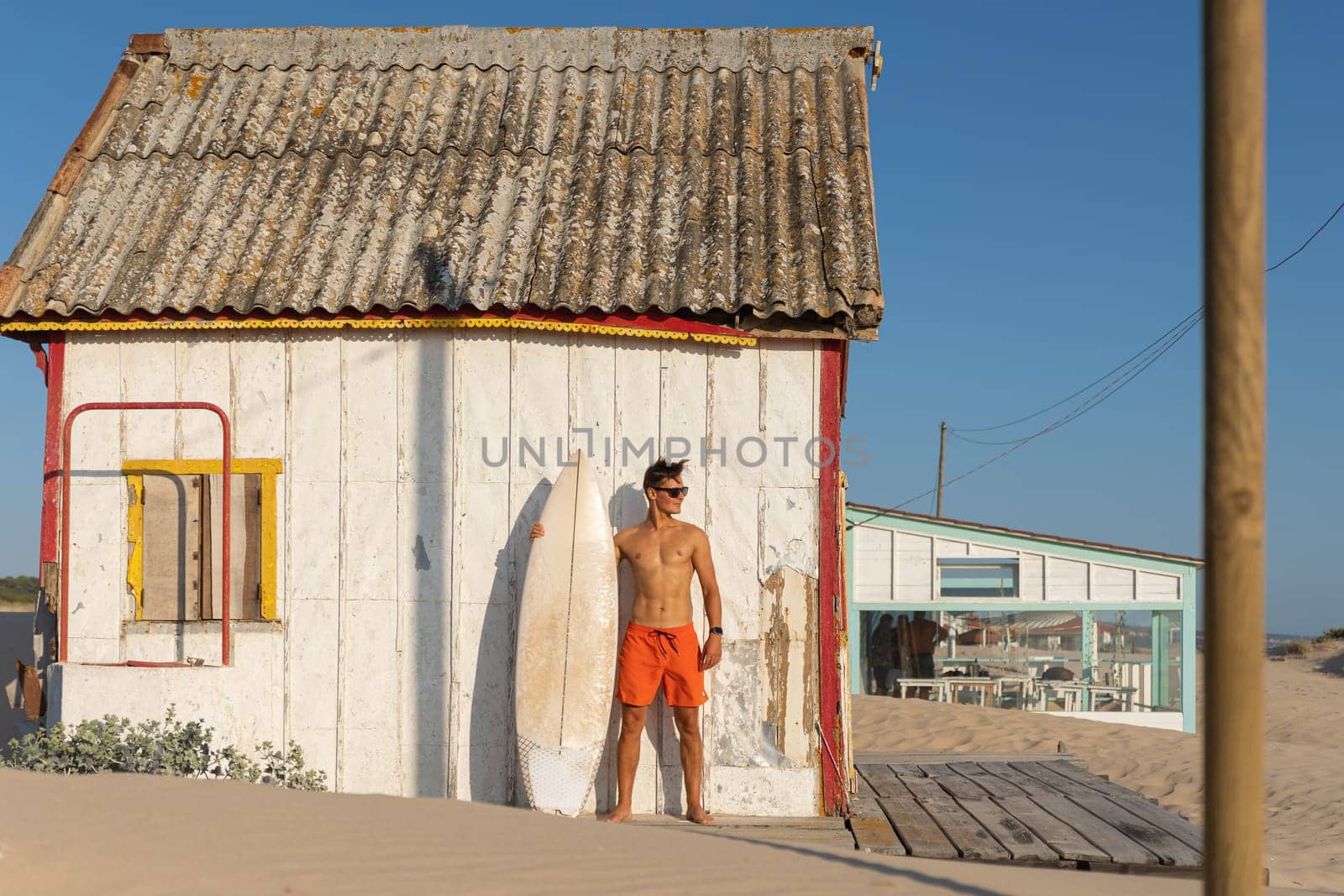 A man with nice athletic body standing at the shore house on the beach with his surfboard - looking to the side. Mid shot