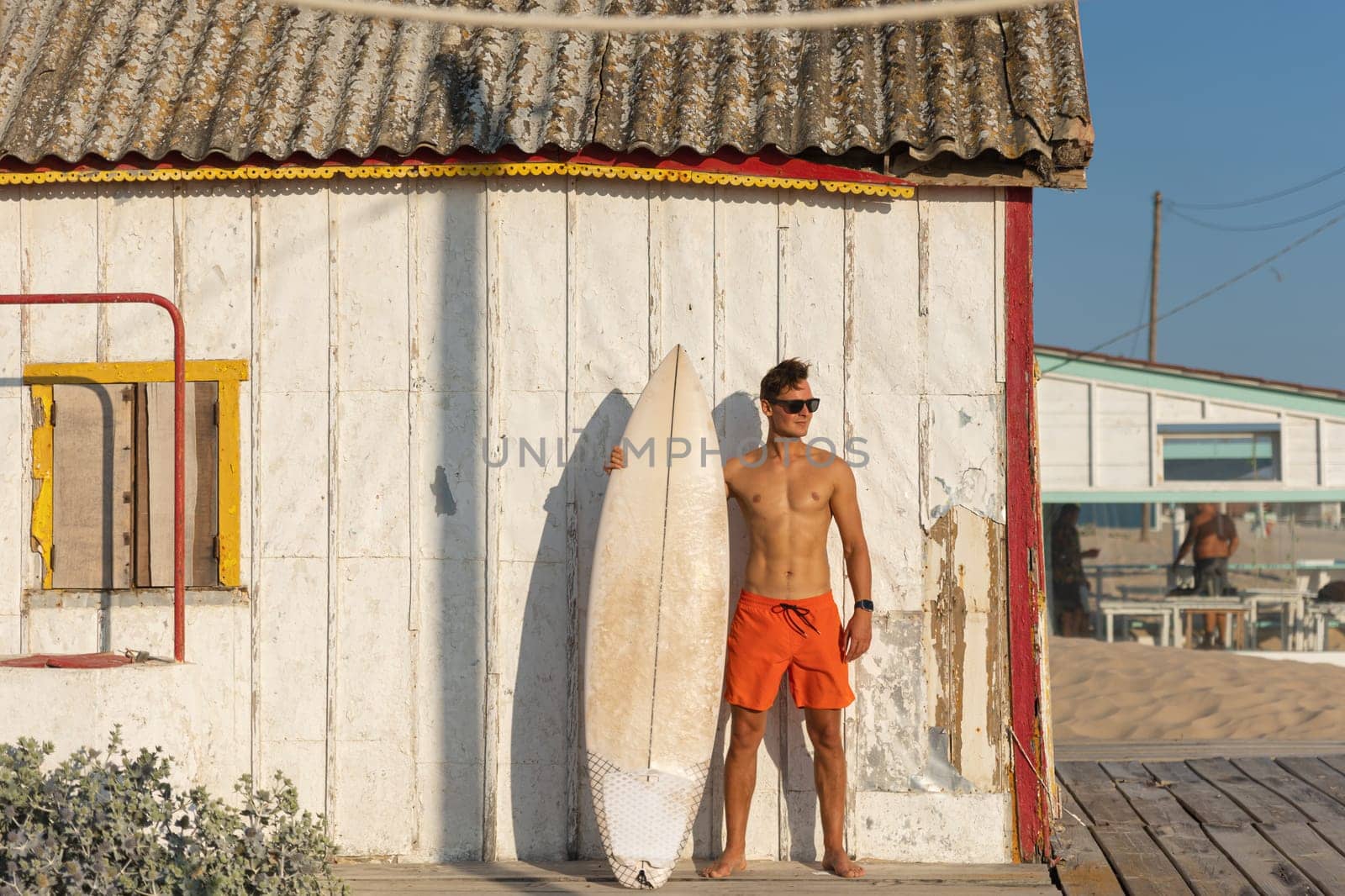 A man with nice athletic body wearing sunglasses standing at the shore house on the beach with his surfboard. Mid shot