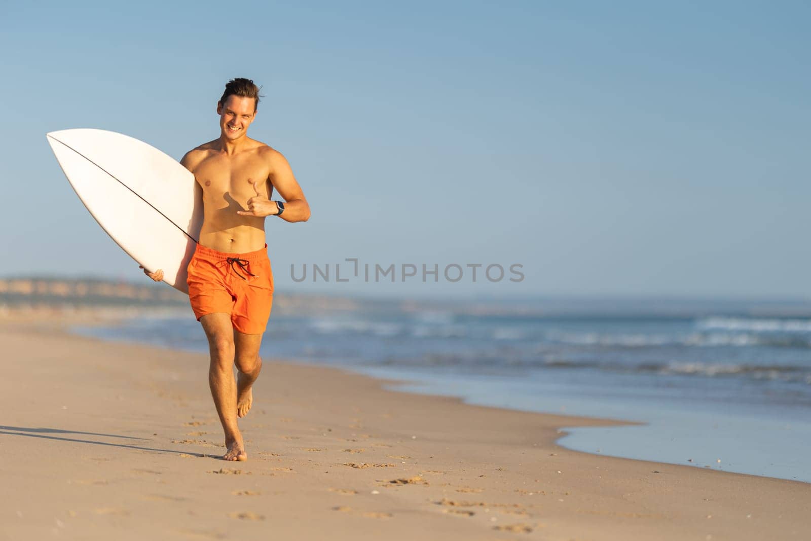 An attractive smiling man with nice athletic body running on the shore holding a surfboard and showing shaka. Mid shot
