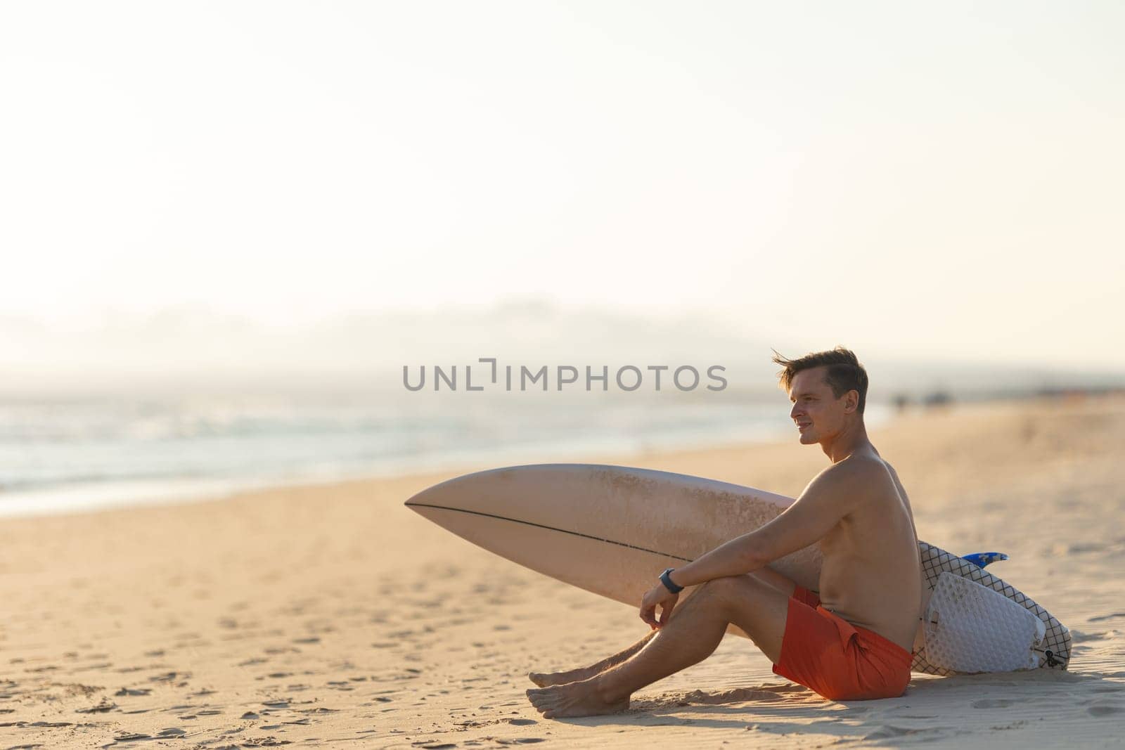 An attractive man with nice athletic body sitting on the shore holding a surfboard. Mid shot
