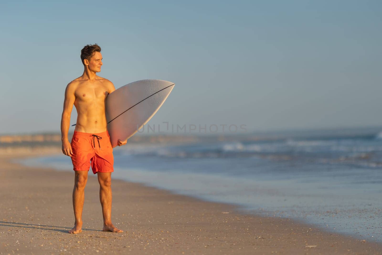 An attractive man with nice athletic body standing on the shore holding a surfboard at early sunset. Mid shot