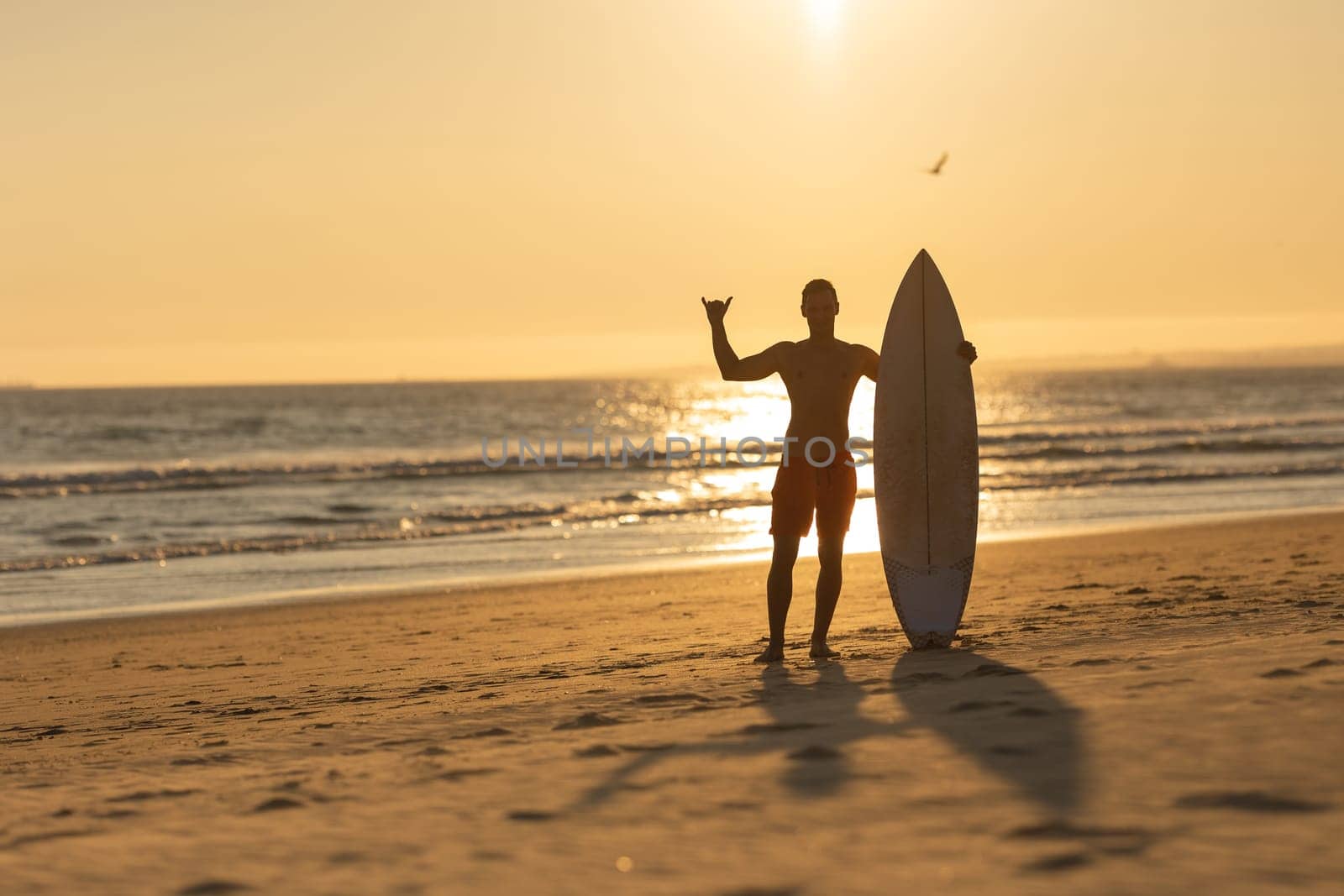 Silhouette of a man standing on the shore holding a surfboard and showing shaka. Mid shot